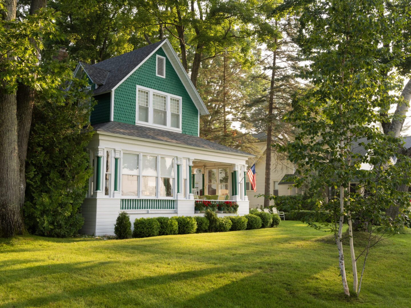 With an American flag proudly displayed, a corner view of a Lake Michigan seasonal summer cottage that is painted green and white. Built in the late 1800's, the house is surrounded by a lush lawn, trees and bushes. It is the perfect escape for vacation, rest and relaxation.